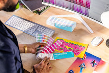 Load image into Gallery viewer, A person at a desk holding a Color Bridge colour guide fan deck, with a matching Color Bridge box and other colour swatch cards on a wooden desk near a computer monitor.