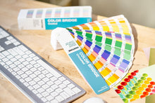 Load image into Gallery viewer, Pantone Color Bridge Guide GG6102 fanned-out colour swatch book lies on a wooden desk next to a white keyboard and mouse, with its box in the background.
