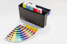 Load image into Gallery viewer, A Pentone Formula Guide fan deck and matching color organizer box on a clean white background. The dark charcoal organizer contains multiple color chips, and the guide in front displays a spectrum of color swatches.
