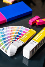 Load image into Gallery viewer, Both Pantone Formula Guide GP1601C solid colour guide books arranged on a glossy dark surface, including a fanned-out guide and one closed volume with bright pink and blue objects on desk.