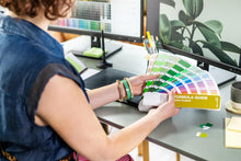 Load image into Gallery viewer, A person seated at a desk holds an open Pantone Formula Guide GP1601C. The scene includes computer monitors, pens, and a plant in an office-like environment.