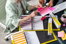 Load image into Gallery viewer, A close-up overhead view of a desk with two people examining colour sample strips and books. Visible items include pink and orange colour cards, binder rings, boxes labelled colour guide, sticky notes, a keyboard, and a coffee cup on a dark desk.